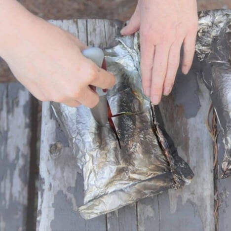 A pair of white hands carefully opening a hot, charred foil packet on a gray picnic table.