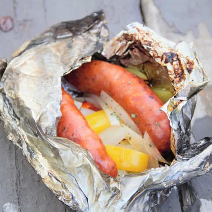 A foil packet with two sausage links, yellow bell pepper strips, and yellow onion slices sits on a gray background.