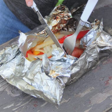 A person holding a knife and fork cutting into an open foil packet stuffed with sausage, peppers, and onions.
