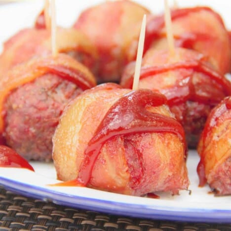 Close up of a Cheese Stuffed Bacon Wrapped Meatball on a serving plate with a toothpick in it, drizzled with sauce, and other meatballs in the background.