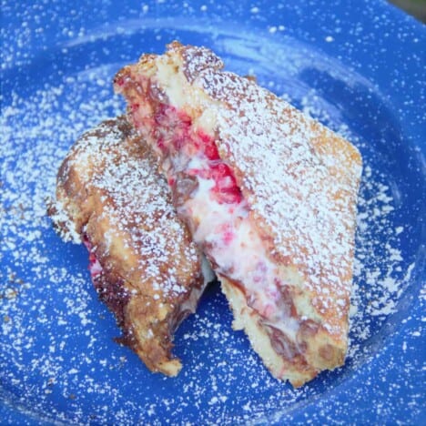 A finished toastie, sitting on a blue camping plate, is cut in half so that the ricotta cheese and raspberries are oozing out.