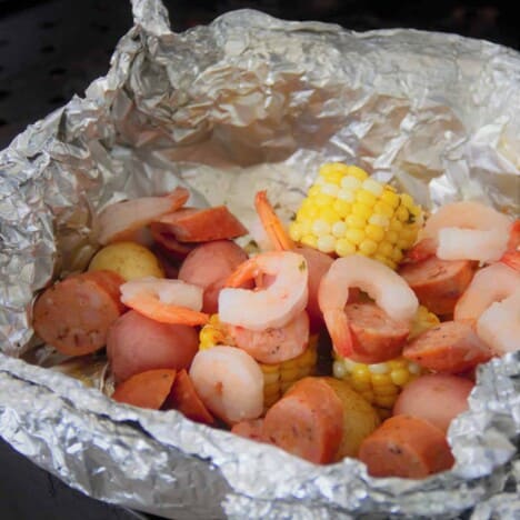A wide shot of a foil packet with shrimp, slices of andouille sausage, cooked baby red potatoes, and chunks of corn on the cob.