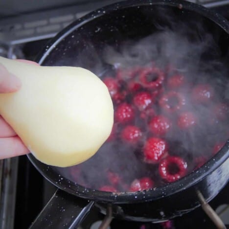 A black camp pot simmering with red wine and raspberries, with a peeled pear half being added.