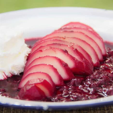 A white camp plate with a sliced pear sitting in a bed of red wine and raspberry sauce and a side of whipped cream.