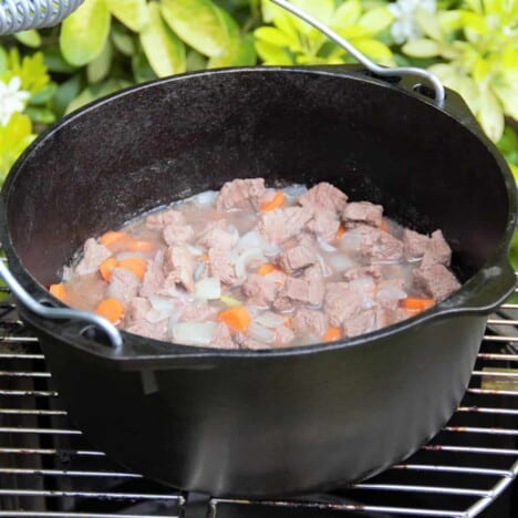 A mixture of lamb, onions, and carrots fills a Dutch oven placed on a grill grate.