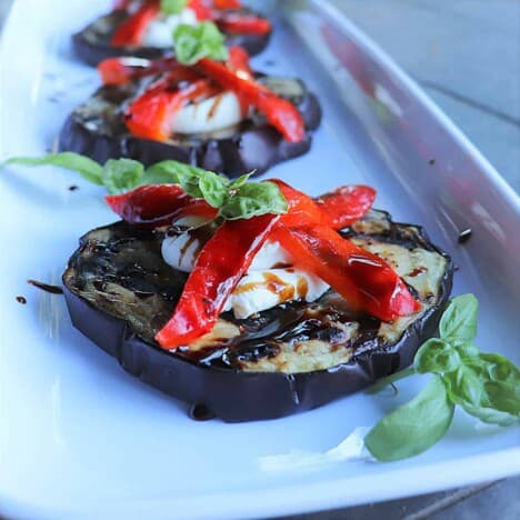 Three stakes of grilled eggplant, topped with goat cheese, red bell pepper slices, basil leaves, and balsamic glaze, sit on a long white serving platter.