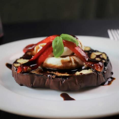 A grilled slice of eggplant, topped with goat cheese, red bell pepper slices, basil, and balsamic vinegar, sits on a white plate.