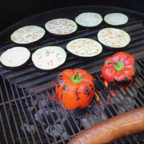 Looking down on to raw eggplant slices and whole red bell peppers starting to cook on a grill with red hot coals.