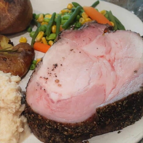 A large slice of grilled herb crusted pork loin sits on a white plate next to mixed vegetables and mashed potatoes.