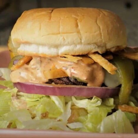 A close up shot of a portobello mushroom burger with layers of shredded lettuce, red onion, sauce, fried tortilla strips, and a bun.