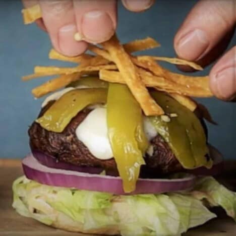 A portobello mushroom burger is layered on top of a slice or red onion and shredded lettuce and being topped with fried tortilla strips.