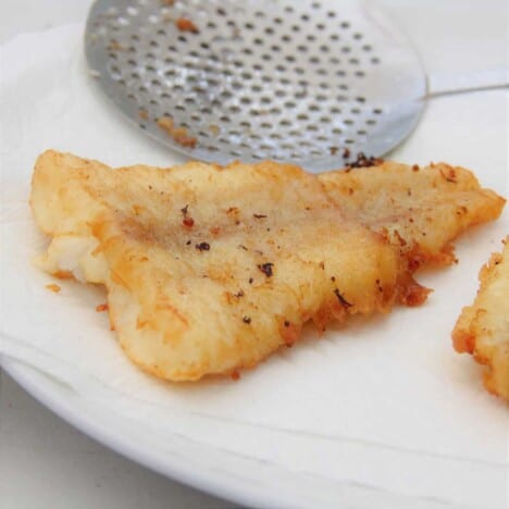 A close up shot of a piece of beer battered fish resting on white paper towels with a slotted spoon in the background.