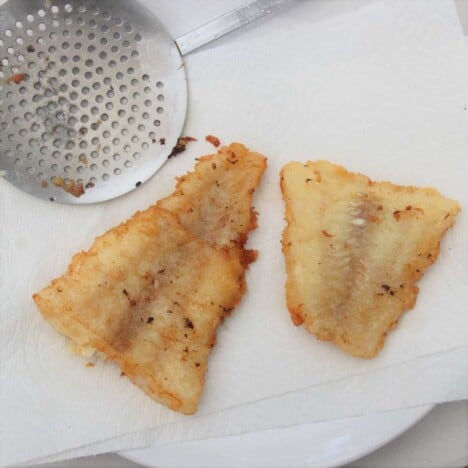 Two fried pieces of beer battered fish lay on paper towels next to a slotted spoon.
