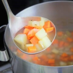 A ladle holding chunks of carrots and potatoes in broth is held above a stock pot.