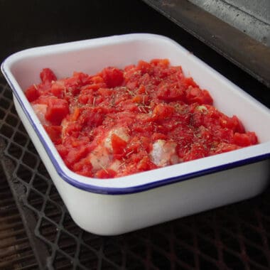 A large white baking dish with blue trim is filled with uncooked chicken drumsticks and diced tomatoes.