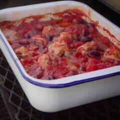 Looking across a large white baking dish with blue trim filled with cooked chicken drumsticks simmered in diced tomatoes with black olives.