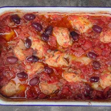 Looking down into a baking dish of chicken thighs simmering in tomato sauce with black olives.