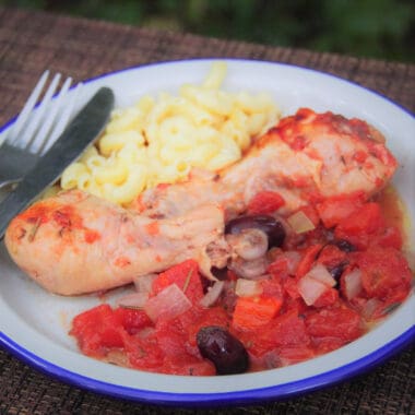 A white plate on a wood background is filled with chicken cacciatore with tomatoes and black olives, with a side of rice.