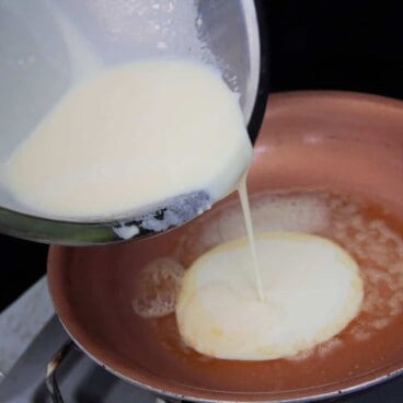 Batter in a metal bowl is being poured into a light brown skillet on a gas stove top.