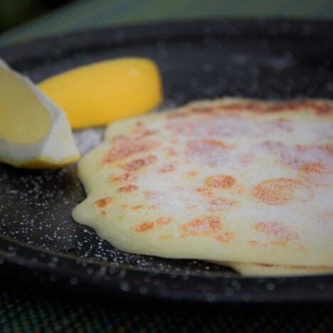 An English pancake sits on a black tin plate with a slice of lemon in the background.