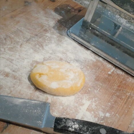 A round of homemade pasta dough sits on a lightly floured wooden work top, with a knife and pasta machine in the background.