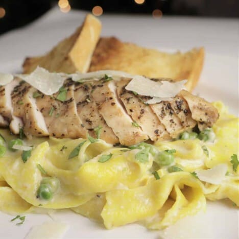A grilled, sliced chicken breast sits atop creamy, golden yellow pasta with green peas, on a white plate.