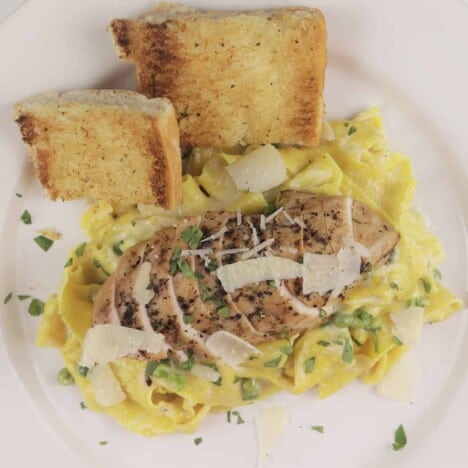 Looking down onto a white plate with a grilled, sliced chicken breast sits atop creamy, golden yellow pasta with green peas.