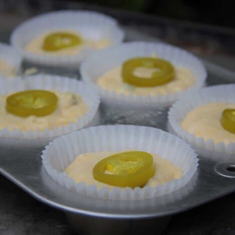 Jalapeno muffin batter with a slice of jalapeno pepper, in a muffin tin lined with white paper liners.