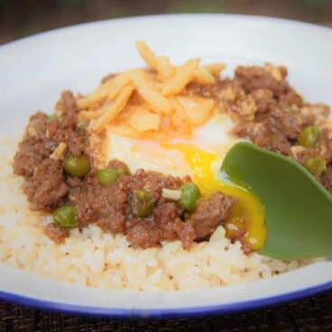 A camp plate with a base of rice topped with ground game stew, an egg, and garnished with fried onion.