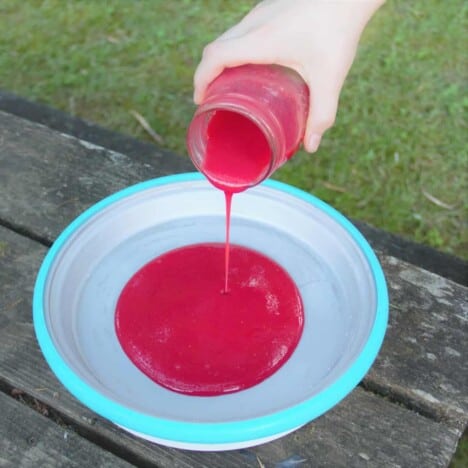 A white hand pours the raspberry puree from a glass jar into a white bowl with a blue rim resting on a picnic table.