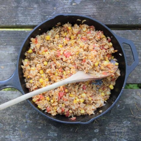 A cast iron skillet, filled with rice, tomatoes, and corn, is resting on a wooden picnic table.