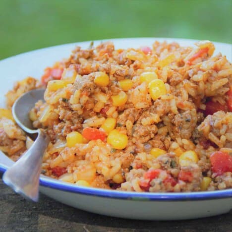 Looking across a shallow white dish filled with rice with chunks of corn and tomatoes with grass in the background.