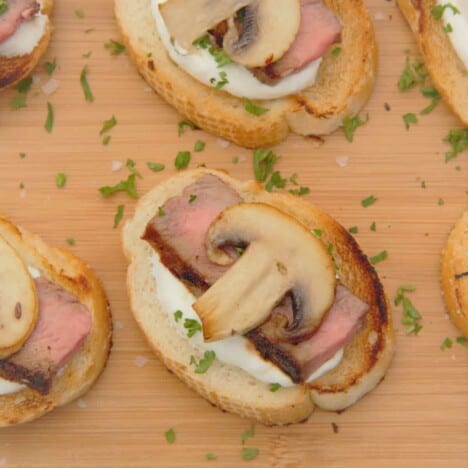 Several crostini with horseradish cream, medium-rare ribeye slices, sliced mushrooms, and chives sit on a wooden cutting board.