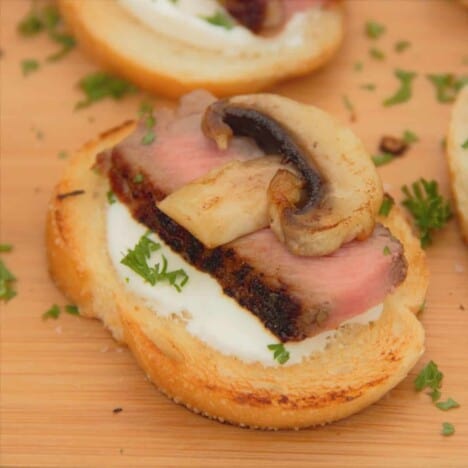 A single crostini, topped with horseradish cream, medium-rare steak, a golden brown mushroom, and chives, sits on a wooden cutting board.
