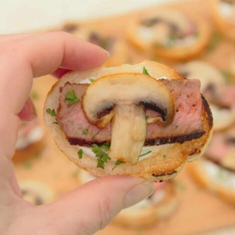 A white hand holds a single crostini, topped with horseradish cream, medium-rare steak, a golden brown mushroom, and chives.