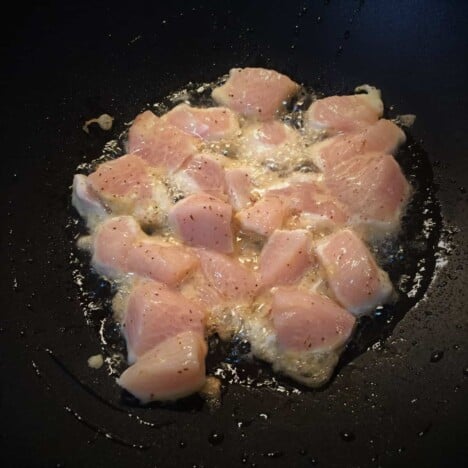 Looking into a dark wok with raw chunks of chicken cooking.