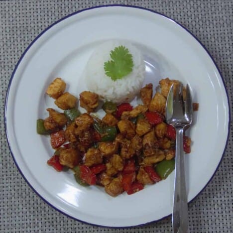 Looking down on a white plate with a blue rim holding a pile of stir fried chicken and bell peppers with a side of rice.