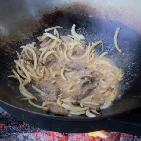 Looking down into a black wok over hot coals, with sliced yellow onions and beef.