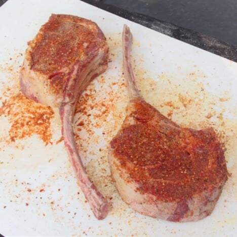 Two tomahawk steaks, placed on a white plate, are liberally sprinkled with BBQ seasoning rub.