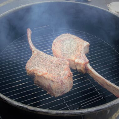 Two raw tomahawk steaks are on a small grill with smoke arising from the cooking meat.