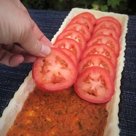 A hand layers slices of tomatoes in a pastry-lined tart tin with a layer of red pesto sauce.