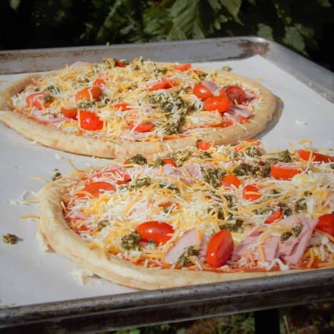 Two uncooked bacon, tomato, and cheese pizzas on a baking sheet with parchment paper.