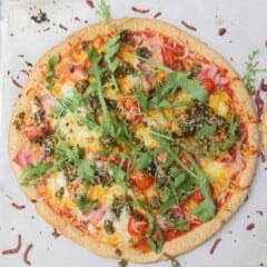 A round bacon, tomato, and cheese pizza, topped with fresh arugula on a white background.