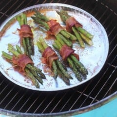 Several bundles of bacon-wrapped asparagus spears are on a sliver serving platter on top of a grill.