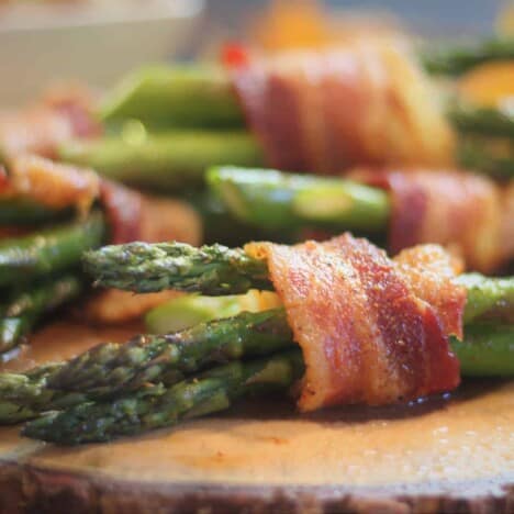 A close up shot of asparagus spears wrapped in cooked bacon, resting on a wooden cutting board.