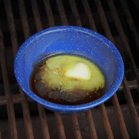 A mixture of melted butter and barbecue sauce sits in a blue bowl on a grill grate.