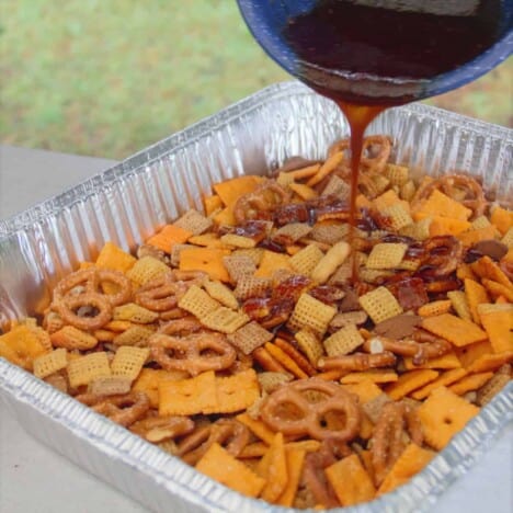 Barbecue party mix is in a foil tray being drizzled with barbecue sauce from a blue bowl.