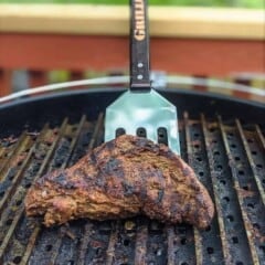 A grilled bavette steak is on a grill and is being lifted by a metal spatula.