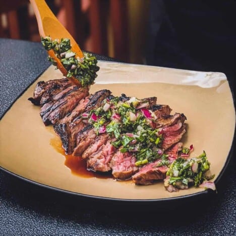A sliced bavette steak is topped with bright green chimichurri sauce and resting on a beige plate on a dark background.