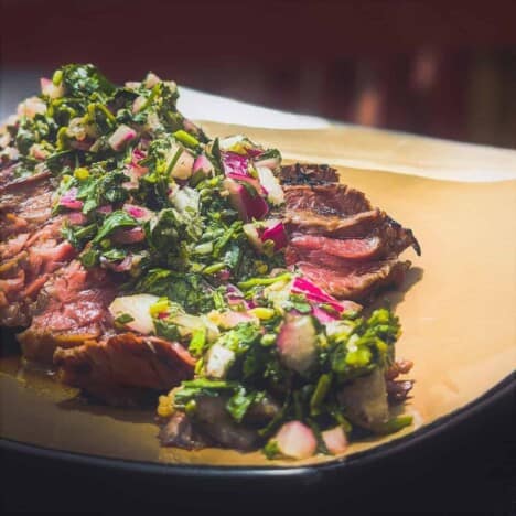 A close up shot of medium-rare grilled bavette steak topped with bright green chimichurri sauce on a beige plate.